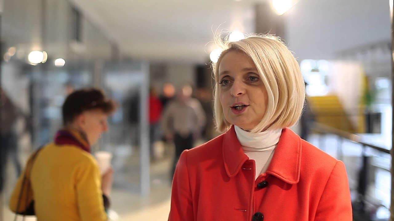 A kép egy női beszélgetést mutat egy nyilvános helyen, háttérben más emberek és modern épületek láth.
