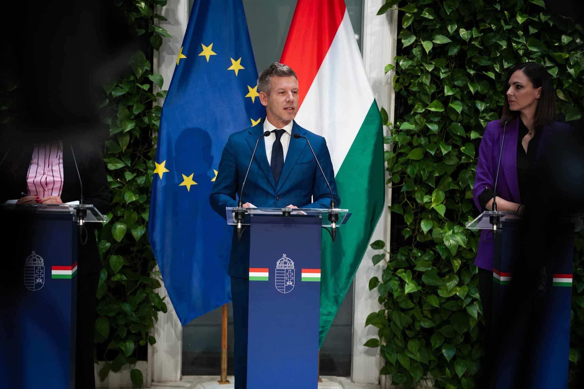 EU zászló és magyar zászló a háttérben, politikai sajtótájékoztató.