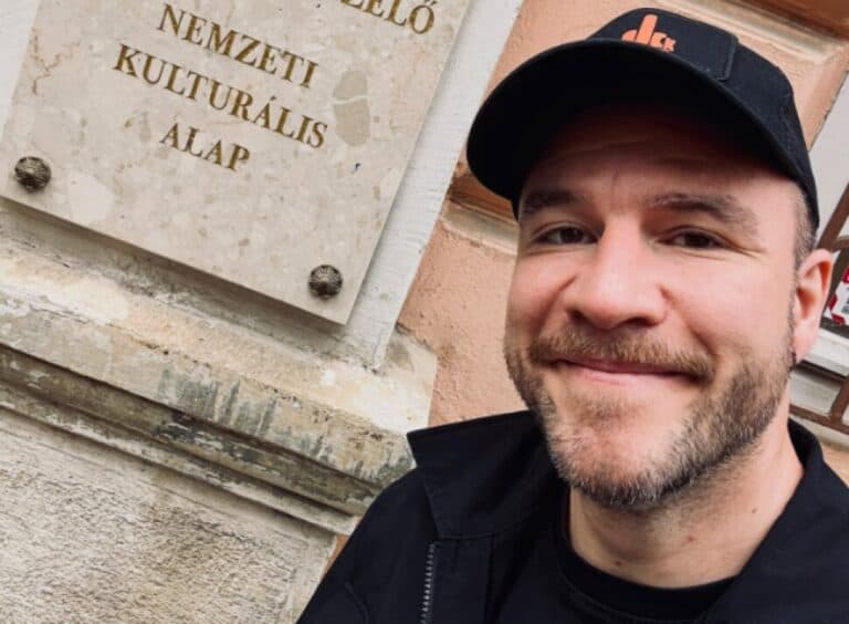 Fiatal férfi mosolyogva áll egy épület előtt, háttérben felirat a Nemzeti Kulturális Alapró.