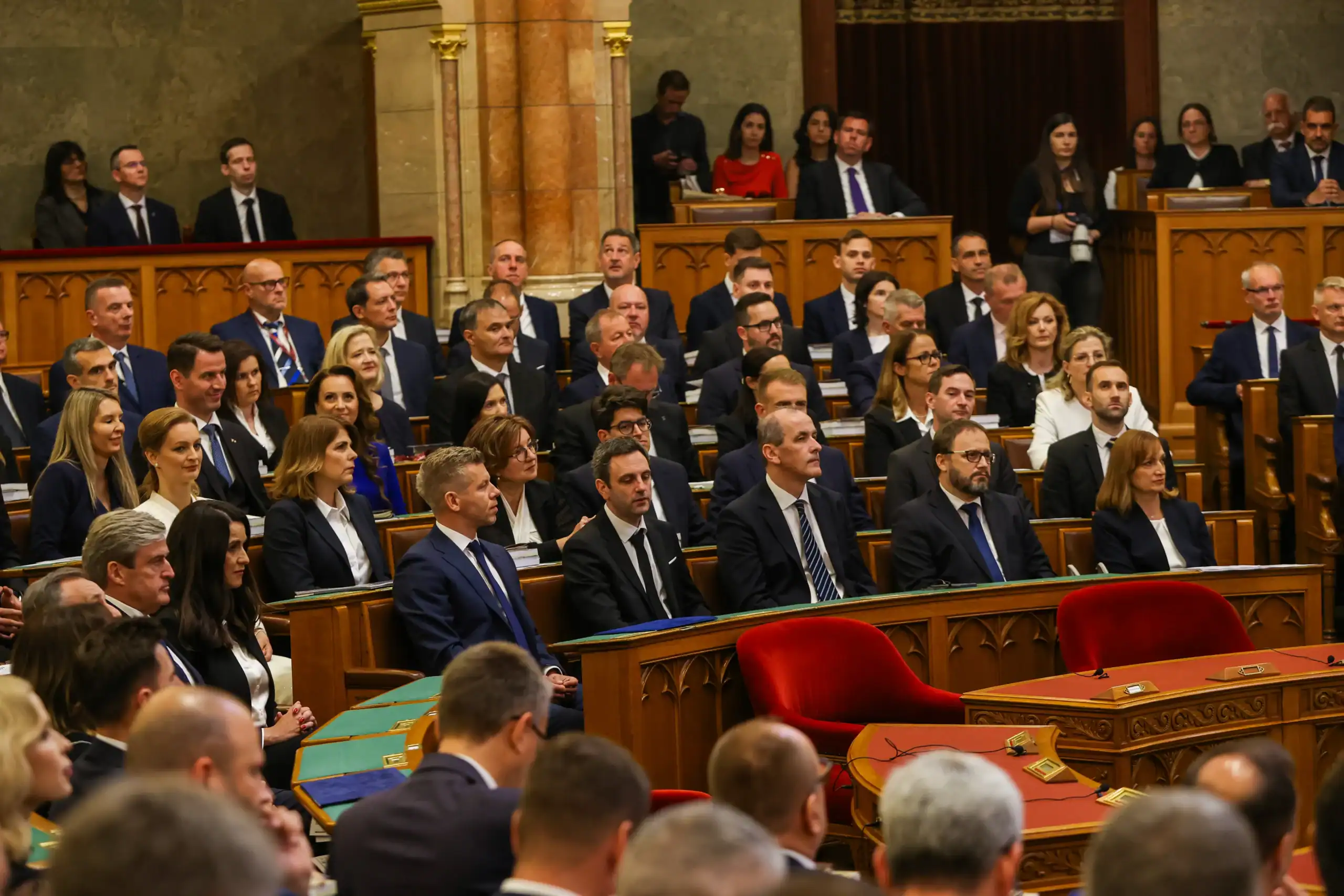 Politikai ülés a parlamentben, ahol képviselők és politikusok hallgatnak.
