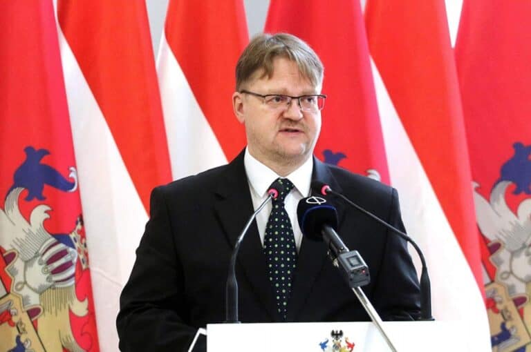 Magyar politikus beszédet mond mikrofon előtt, nemzeti zászlók mögött.