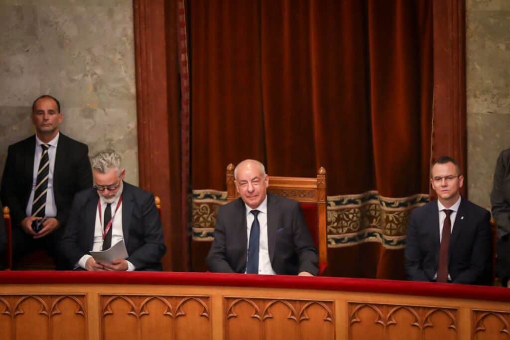 Magyar politikusok ülnek egy hivatalos eseményen, háttérben vörös függöny.