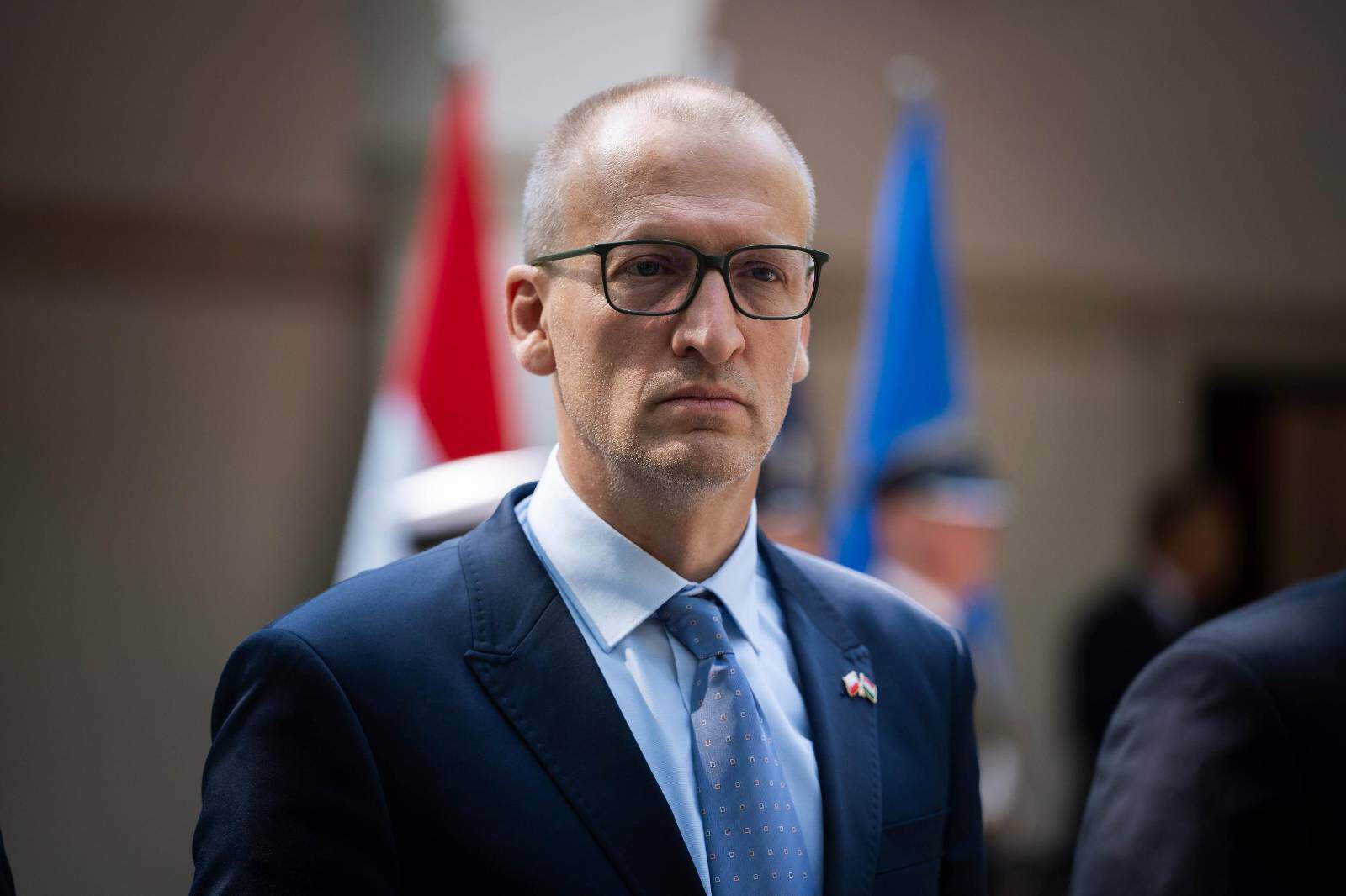 Magyar politikus férfi arcképe, hivatalos eseményen, kék öltönyben és szemüvegben.
