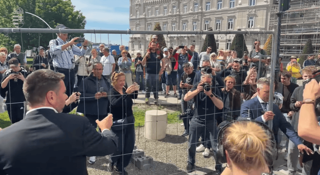 A kép egy nyilvános eseményt vagy tüntetést ábrázol, ahol sokan gyűltek össze, és egy férfi beszédet.
