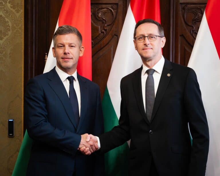 Két férfi kezet fog egy hivatalos eseményen, magyar és nemzetközi zászlók mögött.