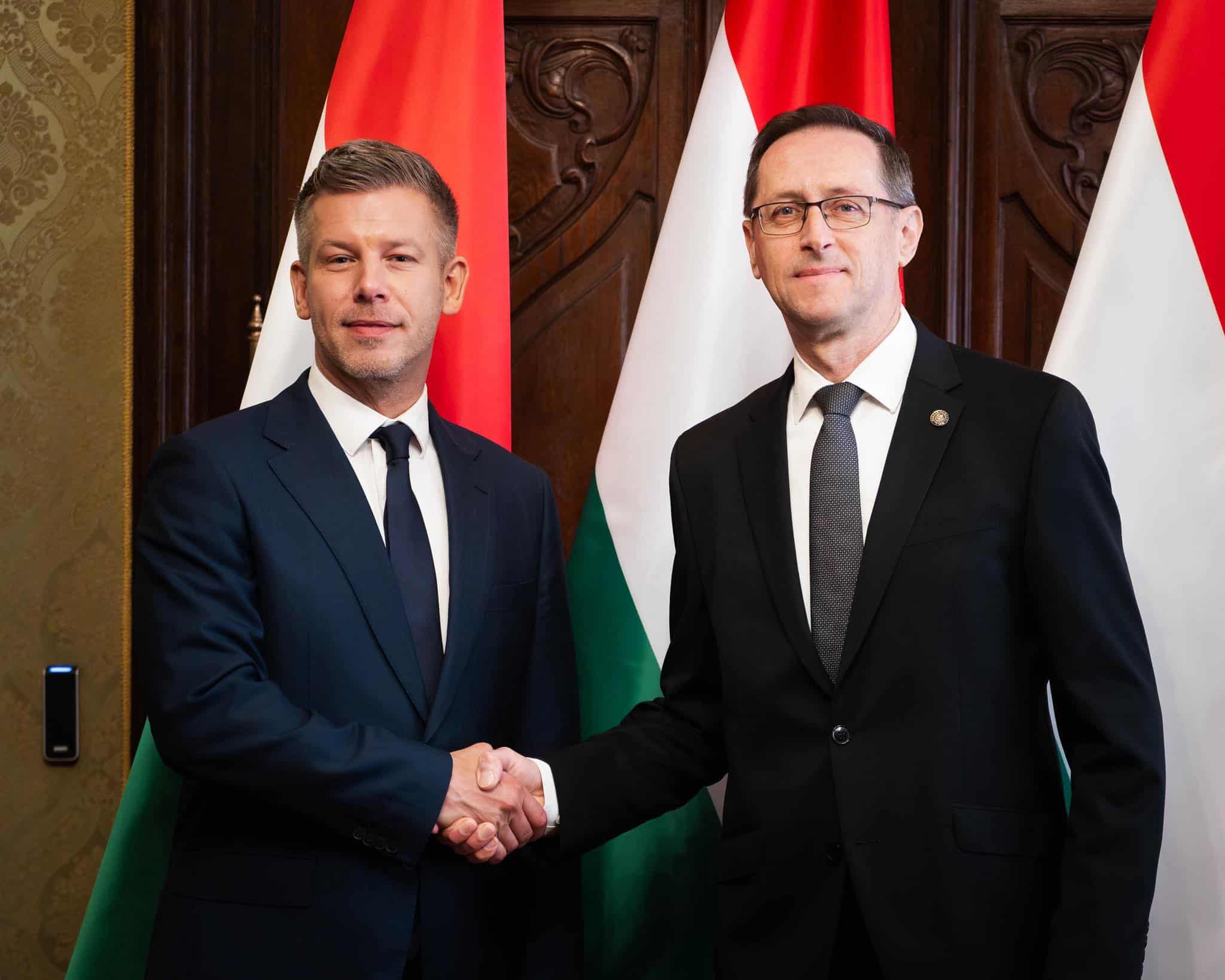 Két férfi kezet fog egy hivatalos eseményen, magyar és nemzetközi zászlók mögött.