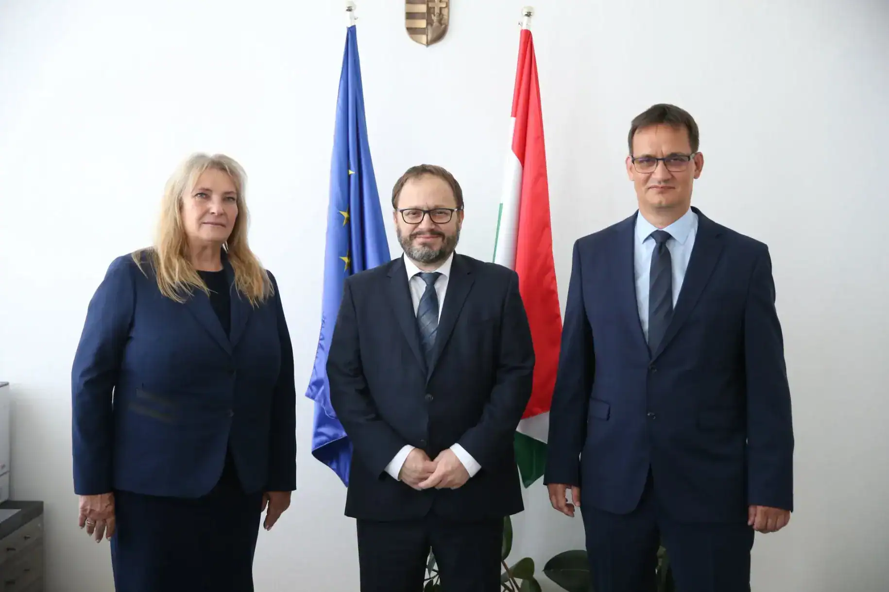 Három személy áll egy irodában, mögöttük az EU és magyar zászlók.
