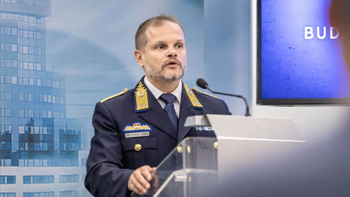 Magyar katonai vezető beszédet tart egy konferencián, háttérben modern irodaépületek.