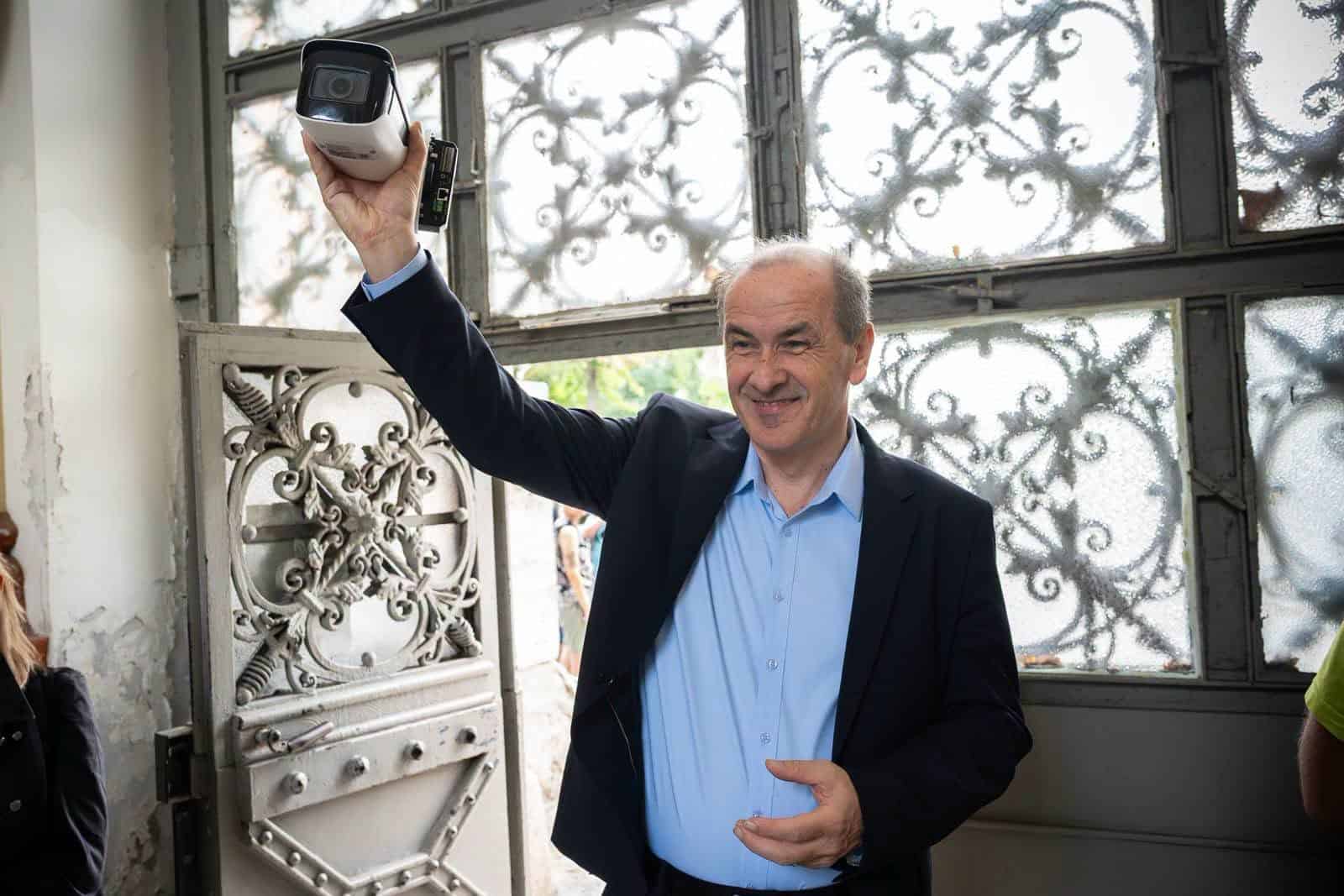 Egy férfi fényképezőgéppel a kezében mosolyogva áll egy nyitott ajtó előtt.