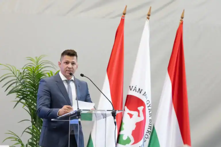Lőrincz Tamás beszédet mond egy konferencián, háttérben magyar zászlók.