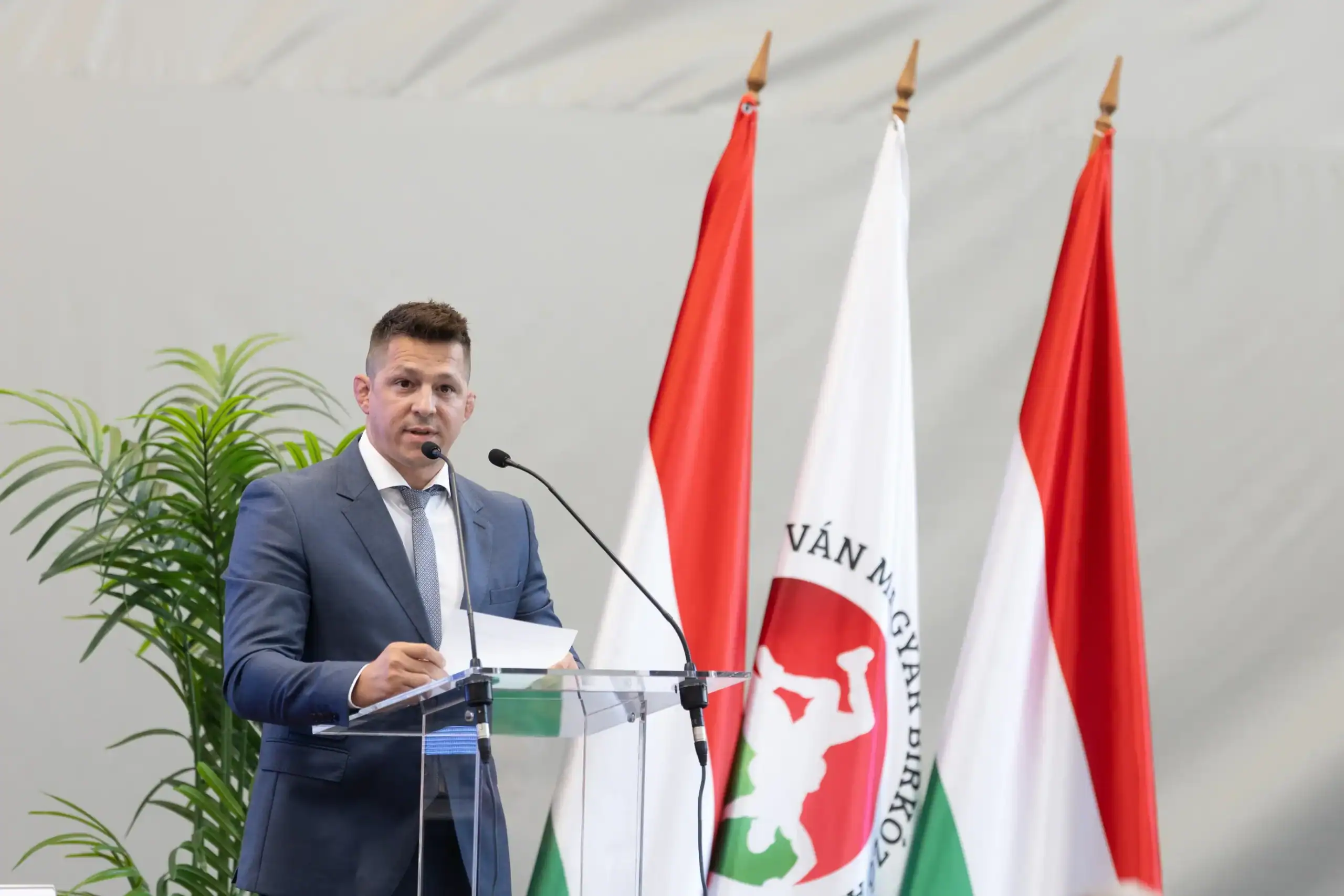 Lőrincz Tamás beszédet mond egy konferencián, háttérben magyar zászlók.
