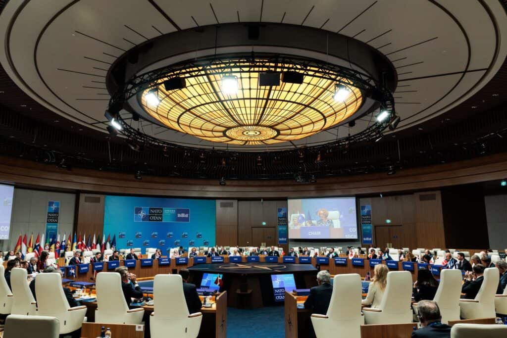 Az NATO csúcstalálkozó nagyterme, körülötte ülő diplomaták és képviselők.