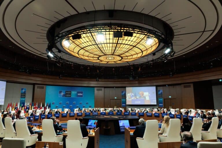 Az NATO csúcstalálkozó nagyterme, körülötte ülő diplomaták és képviselők.
